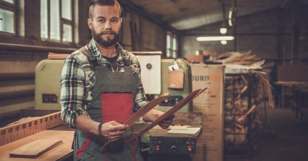 A professional woodworker is in his workshop next to a bench and holding up two scraps of wood for comparison.