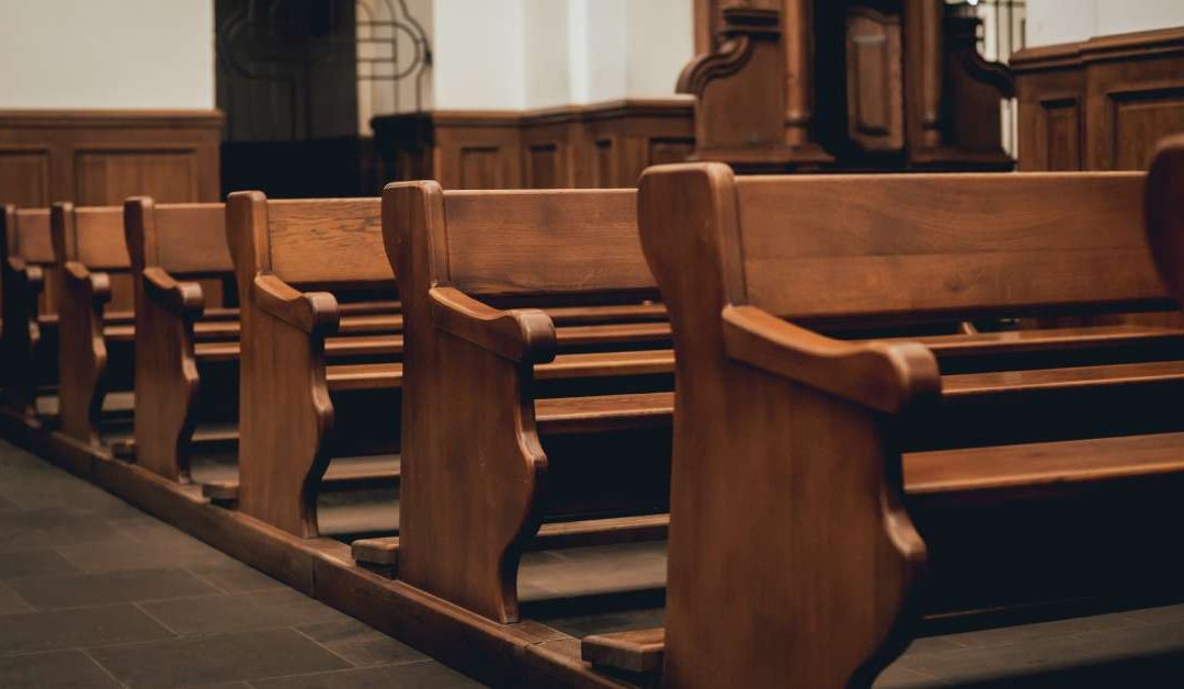 A row of pews made of hard wood is lined up in a church with brick flooring and beige walls. A pillar is in the background.