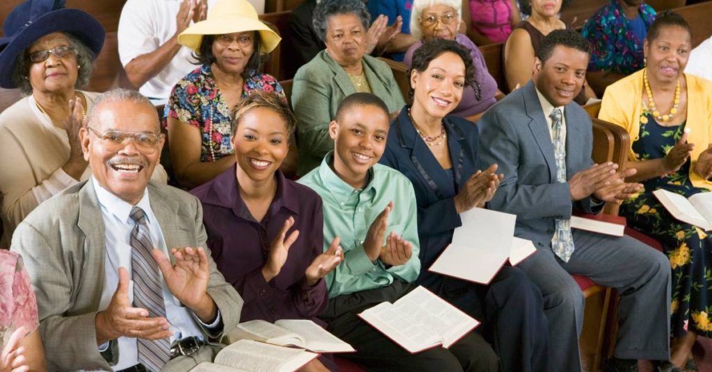 A congregation dressed in formal clothing smiling at the camera while clapping during service in a church.