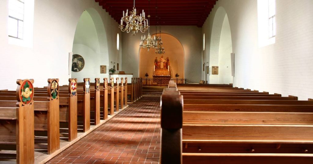 The interior of a church with rows of pews on either side of the aisle. There are chandeliers hanging on the ceiling.