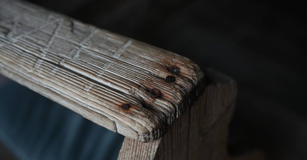 A closeup of the arm of a church pew with old split wood and rusty screws in need of a deep restoration.