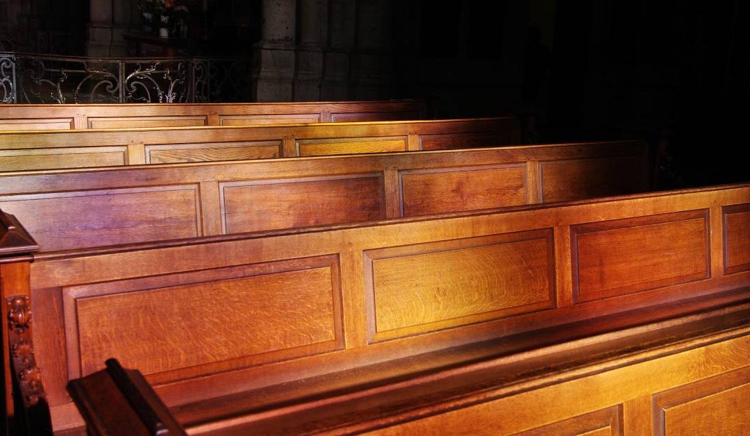 A row of wooden church pews in an old church with a small amount of light streaming into the room and shining on them.